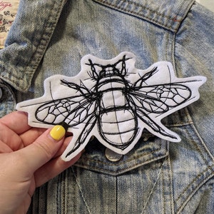 May include: A white and black embroidered bee patch. The patch shows a detailed sketch of a bee with black thread on a white felt background. The patch is attached to a denim jacket.