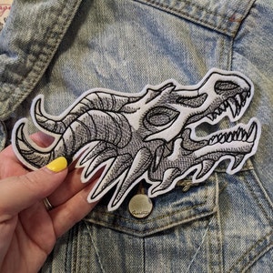May include: A white embroidered patch of a dragon skull with black stitching. The patch is on a blue denim jacket.