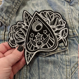 May include: Black and white embroidered patch of a Ouija board with a crescent moon and stars, surrounded by roses.