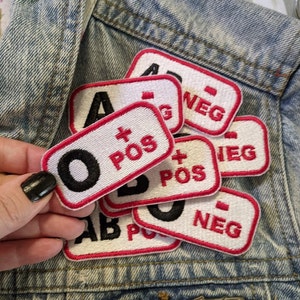 May include: A set of eight embroidered iron-on patches with different blood types: O+, A+, B+, AB+, O-, A-, B-, and AB-. The patches are white with red borders and black lettering.