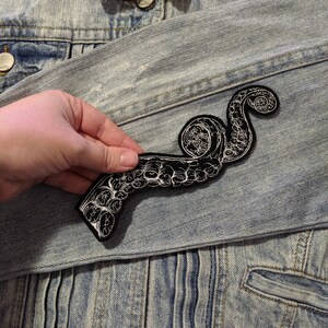 May include: A black and white embroidered octopus tentacle patch. The patch is sewn onto a light blue denim jacket.