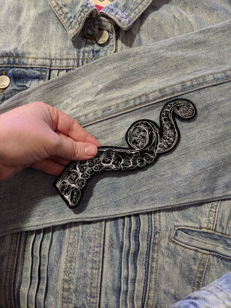 May include: A black and white embroidered octopus tentacle patch. The patch is sewn onto a light blue denim jacket.