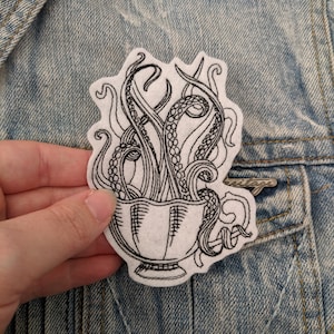 May include: Black and white embroidered patch of an octopus in a cup. The octopus's tentacles are curled up inside the cup.