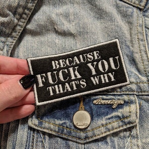 May include: A black rectangular patch with white embroidered text that reads "BECAUSE FUCK YOU THAT'S WHY." The patch is on a denim jacket pocket, next to a silver pin and a round pendant.