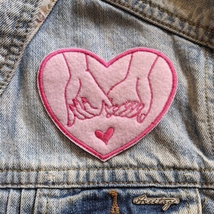 May include: Pink felt heart-shaped patch with two hands holding each other inside the heart. A smaller pink heart is in the center of the larger heart.
