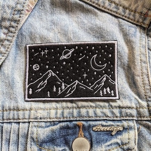 May include: A black embroidered patch with a white outline of a mountain range under a starry night sky. The patch features a crescent moon, a planet with rings, and a shooting star.