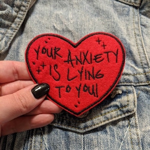 May include: Red felt heart-shaped patch with black embroidered text that reads "Your anxiety is lying to you".