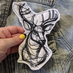 May include: A black and white embroidered patch of a deer head with intricate line work. The patch is on a denim jacket.