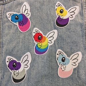 May include: Five embroidered iron-on patches featuring cartoon bees with different coloured stripes. The bees are all in the same pose, with their wings spread and their heads tilted to the side. The patches are all on a blue denim background.