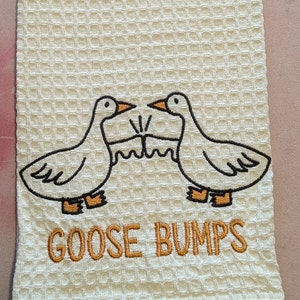 K&ouml;nnte beinhalten: Wei&szlig;es K&uuml;chentuch mit gestickten G&auml;nsen und dem Text "Goose Bumps".