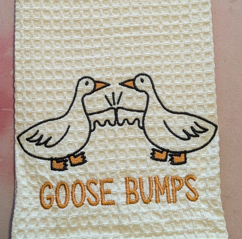 K&ouml;nnte beinhalten: Wei&szlig;es K&uuml;chentuch mit gestickten G&auml;nsen und dem Text "Goose Bumps".