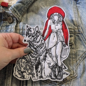 May include: Embroidered iron-on patch featuring a girl in a red hooded cloak and a wolf. The girl is wearing a white dress and boots. The wolf is sitting next to her and looking to the left. The patch is on a denim background.