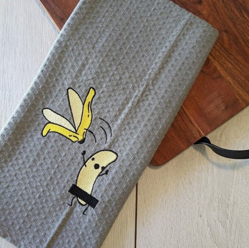 May include: Gray waffle-weave kitchen towel with an embroidered banana design. The design features a peeled banana and a banana with a censor bar. The towel is on a wooden cutting board.