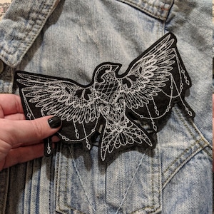 May include: Black embroidered iron-on patch of a bird with white thread, with dangling chains on the bottom.