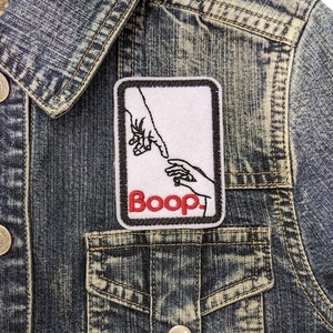 May include: A white embroidered patch with a black outline featuring two hands reaching towards each other. The text "Boop" is in red lettering.