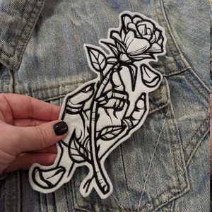 May include: Black and white embroidered iron-on patch of a hand holding a rose.