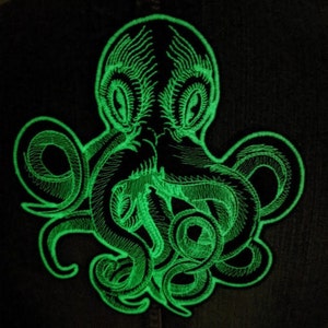 May include: A green glow-in-the-dark embroidered octopus patch. The octopus has eight tentacles and is facing forward with its eyes open.