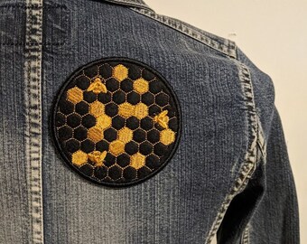 Könnte beinhalten: Ein gestickter Patch in Schwarz und Gold mit einem Wabenmuster und drei Bienen. Der Patch ist auf eine blaue Jeansjacke genäht.