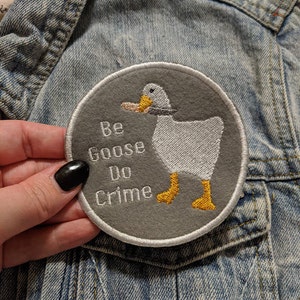 May include: A grey, round patch with white stitching. The patch features a white goose with yellow feet and a black eye. The goose is holding a small, black object in its beak. The text "Be Goose Do Crime" is embroidered in white on the patch.