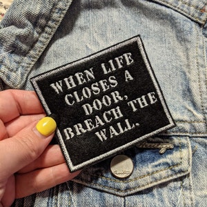 May include: Black and white embroidered patch with the text "When life closes a door, breach the wall."
