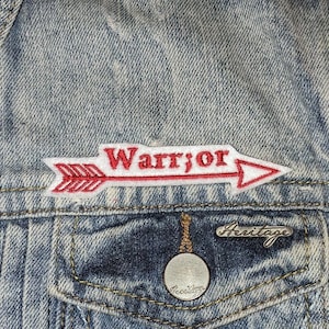 May include: A white patch with a red arrow and the word "Warrior" in red lettering sewn onto a blue denim jacket.