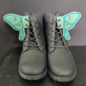 May include: A pair of black lace-up boots featuring decorative mint green moth wing attachments secured by the laces. The wings display detailed dark green vein patterns and small yellow circular accents, contrasting against the dark boot material.