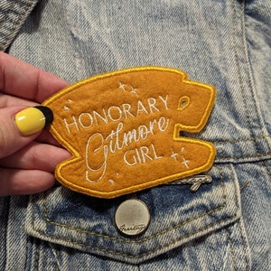 May include: A brown felt patch with yellow stitching, featuring the text "HONORARY Gilmore GIRL" in white embroidery. The patch has a star design on either side of the text.