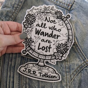 May include: A white embroidered patch with a black outline of a globe with flowers around it. The text "Not all who wander are lost" is embroidered in black on the globe. The text "J.R.R. Tolkien" is embroidered in black at the bottom of the patch.