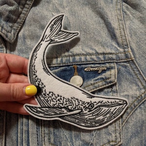 May include: A white embroidered patch of a whale with black outlines on a denim jacket. The whale is facing the right side of the image and has a detailed design.