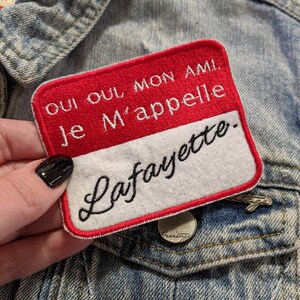 May include: Red and white fabric name tag with the text "Oui oui mon ami. Je m'appelle Lafayette."