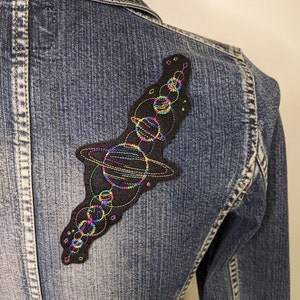 May include: A black denim jacket with a colorful embroidered patch of a solar system on the back. The patch features planets, stars, and rings in rainbow colors.