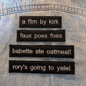 May include: Four rectangular black embroidered patches with white text arranged vertically. The patches read: a film by kirk, faux poes foes, babette ate oatmeal!, and rory's going to yale!. Each patch features a clean, stitched border design.