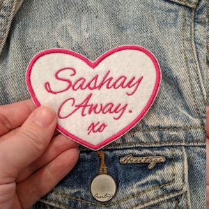 May include: A white heart-shaped patch with pink stitching. The patch says "Sashay Away. xo" in pink cursive lettering.