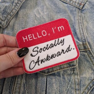 May include: A white and red embroidered patch with the text "HELLO, I'm Socially Awkward." on a denim background.