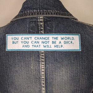 May include: A blue denim jacket with a rectangular patch. The patch has a white background with a light blue border and text that reads: "YOU CAN'T CHANGE THE WORLD. BUT YOU CAN NOT BE A DICK, AND THAT WILL HELP."