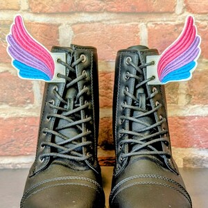 May include: A pair of black combat boots with colorful wing-shaped accessories. The wings are pink, purple, and blue. The boots have laces and a zipper. The background is a brick wall.
