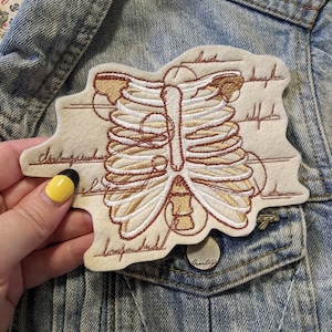 May include: A white felt patch with brown embroidery of a rib cage with anatomical labels. The patch is on a blue denim jacket.