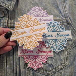 May include: Three embroidered patches featuring a floral design and the text "River Eowyn". The patches are in different colours: gold, blue and pink.