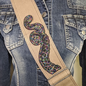 May include: A tan guitar strap with a black iridescent embroidered octopus design. The strap is worn over a denim jacket.