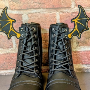 May include: Two black combat boots with black laces, each adorned with a bat wing applique in black and gold. The boots are set against a brick wall background, creating a striking contrast.