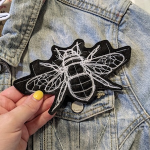 May include: A black embroidered bee patch with white stitching. The patch is on a denim jacket.