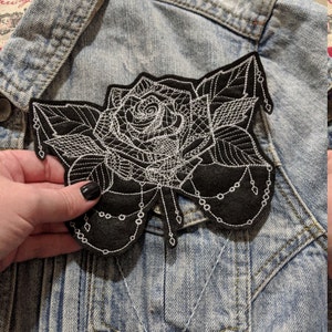 May include: A black felt patch with a white embroidered rose design. The rose has a detailed design with leaves and a chain-like pattern.