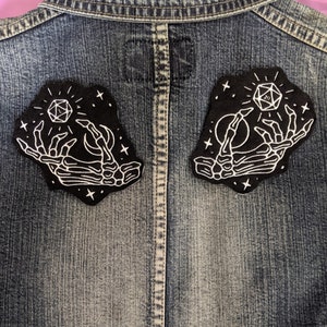 May include: Two black embroidered patches sewn onto a denim jacket. Each patch features a white skeletal hand holding a glowing 20-sided die.