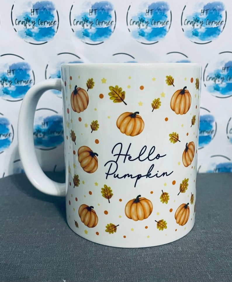 May include: White ceramic mug with a fall-themed design. The mug features a pattern of orange pumpkins, brown leaves, and white stars. The text "Hello Pumpkin" is printed in black on the mug.