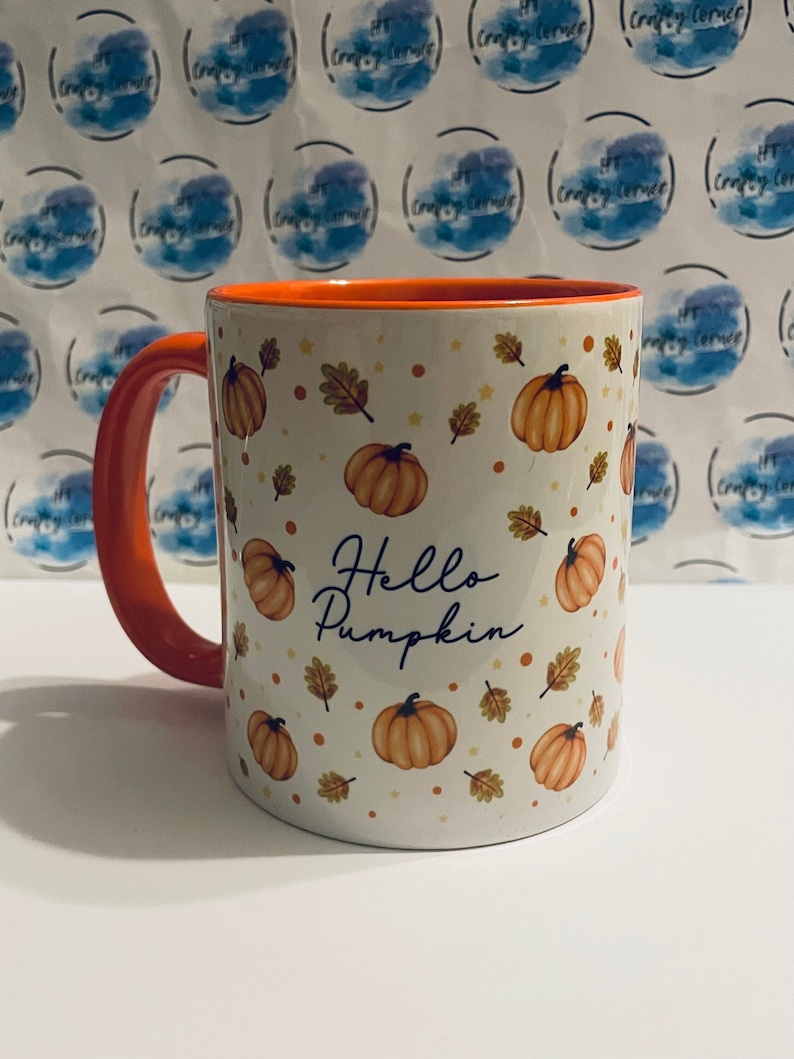 May include: A white ceramic mug with an orange rim and handle. The mug has a pattern of orange pumpkins, brown leaves, and white stars on a white background. The text "Hello Pumpkin" is written in black cursive on the mug.