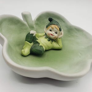 May include: A ceramic leaf-shaped dish with a light green interior and white exterior. A small, whimsical elf figurine in green and yellow clothing reclines inside the dish. The elf has a smiling face and a pointed hat.