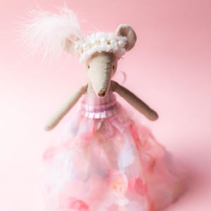 May include: A charming stuffed mouse doll in a pink dress with a layered skirt. The doll has a white floral headpiece with pearls and a feather. The background is a soft pink.