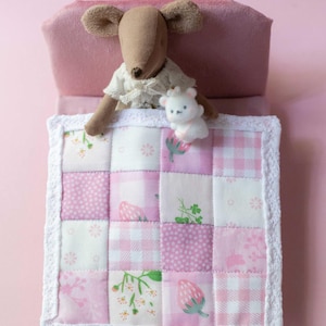 May include: A miniature bed with a pink headboard and a patchwork quilt. A stuffed mouse wearing a white dress and a small white bear are on the bed. The quilt has a white lace border and pink squares with floral and strawberry patterns.