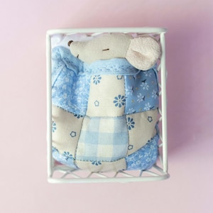 May include: A stuffed mouse toy with a light gray head and pink ears, wearing a blue patchwork quilt. The mouse is inside a white wire cage, set against a pink background. The quilt features floral and gingham patterns.