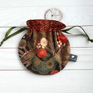 Può includere: Pochette rotonda con coulisse con un design floreale di ispirazione giapponese. La pochette ha una base marrone scuro con motivi floreali rossi, bianchi e dorati. La parte superiore è rosso intenso con un motivo a onde e ha cordini verdi.